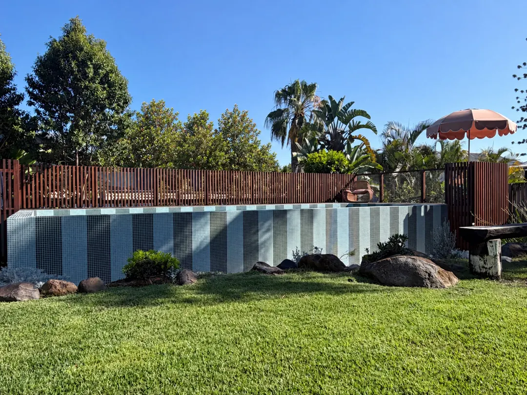 The striped tiled wall of the plunge pool from the lawn, with timber battens, rocks and a scalloped umbrella under a blue sky