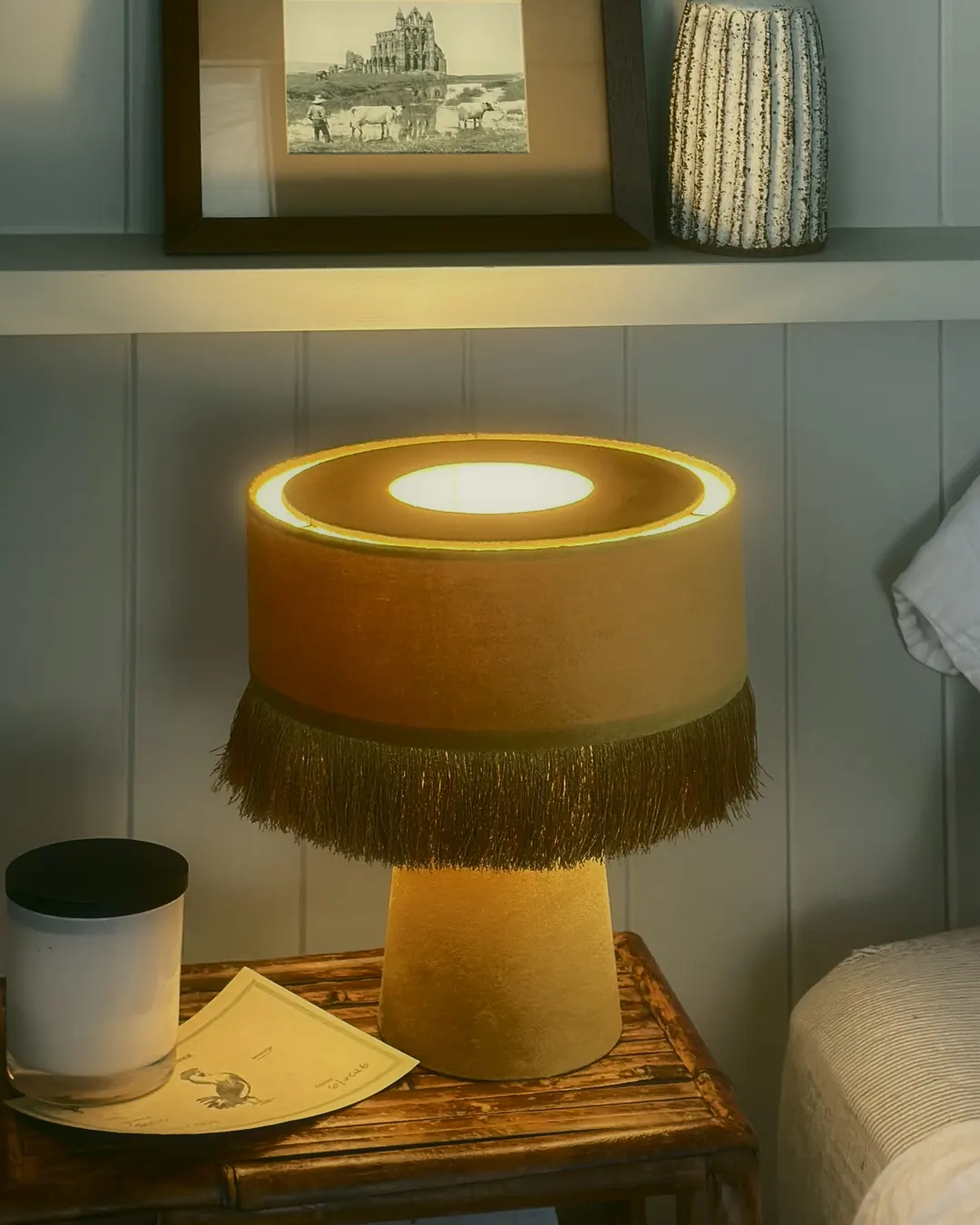 A mustard fringed lamp glowing on a bamboo bedside table, with a candle beside it