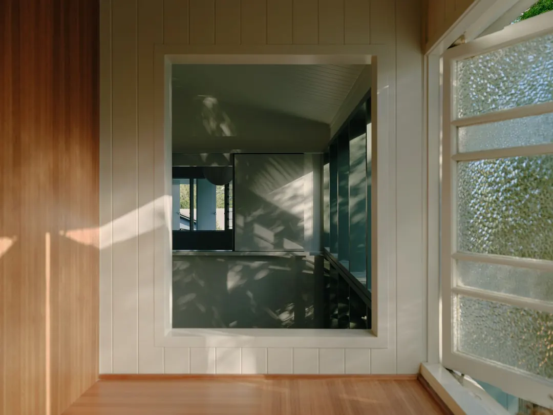 An internal window in a VJ-lined room, framing the stair void, beside an open casement of textured glass